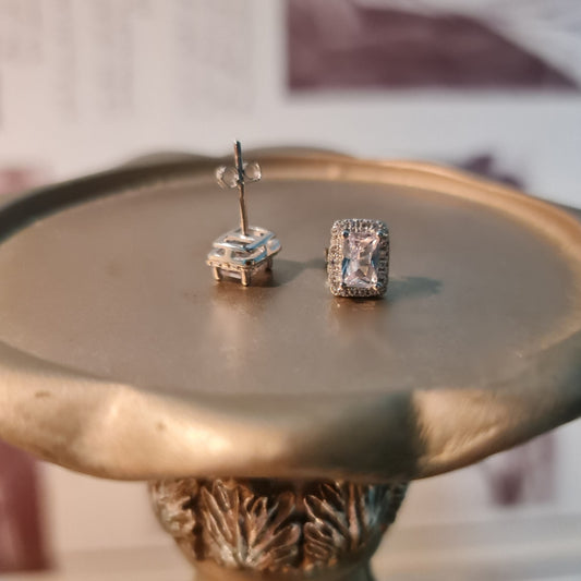 A pair of rectangular silver earrings with zircon stones displayed on a metallic surface with an ornate edge.