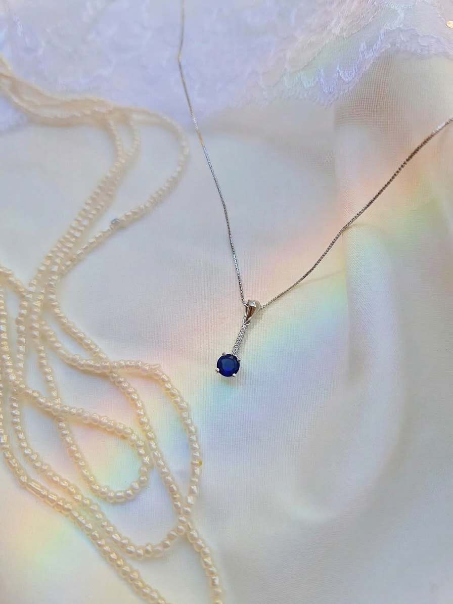 A delicate silver necklace with a single deep blue oval gemstone pendant, displayed on a soft white fabric background with a second strand of cream pearls draped alongside it.