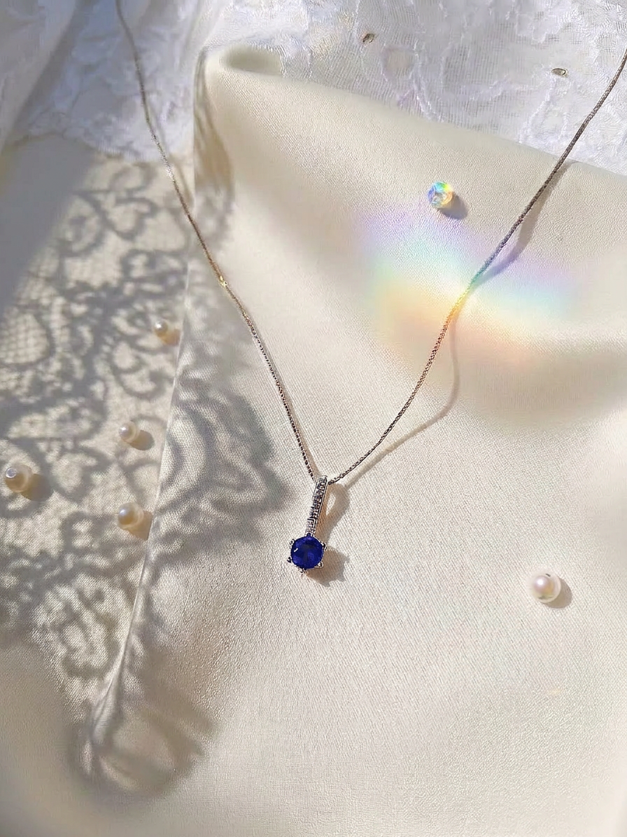 A delicate silver chain necklace with a single deep blue gemstone pendant, displayed on a cream satin fabric with lace shadows and small pearls.