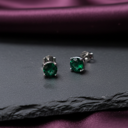 Pair of green gemstone stud earrings on a dark surface with purple fabric background