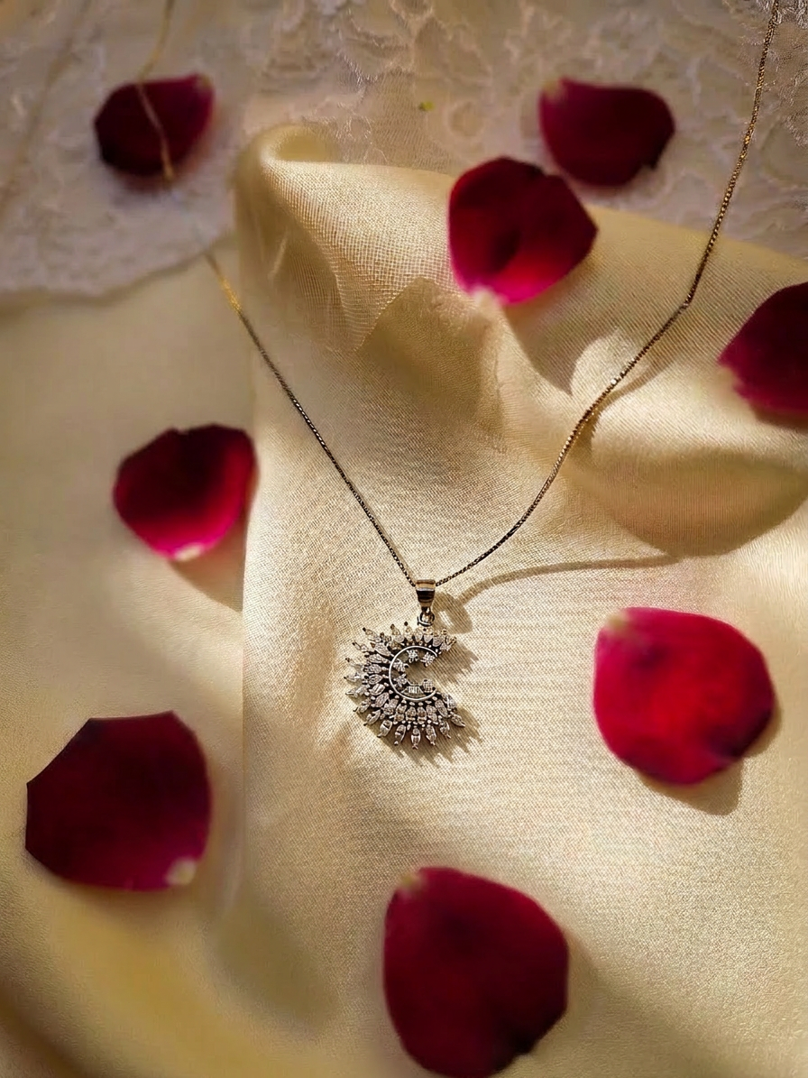A delicate silver-toned necklace with a crescent moon pendant adorned with small clear stones, draped over cream satin fabric scattered with deep red rose petals.