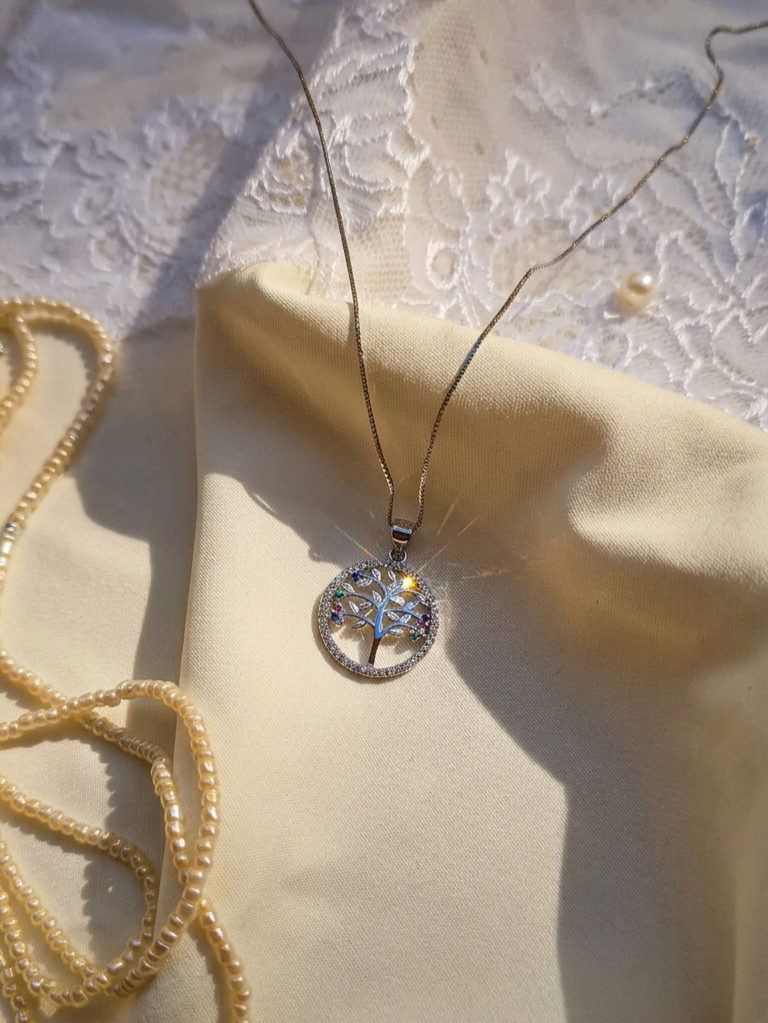 A silver-toned circular pendant necklace with a tree of life design and iridescent blue-green stones, displayed on a cream satin fabric with a pearl necklace and lace in the background.