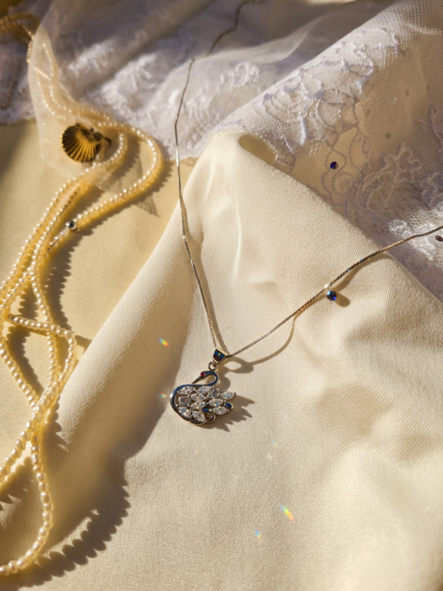 A delicate silver necklace with a swan-shaped pendant adorned with clear crystals, styled on draped cream satin fabric alongside strands of pearl necklace and a small gold shell.