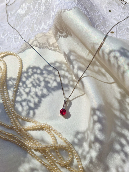 A delicate silver chain necklace with a small red gemstone pendant, styled on draped white satin and lace fabric alongside strands of cream pearls.